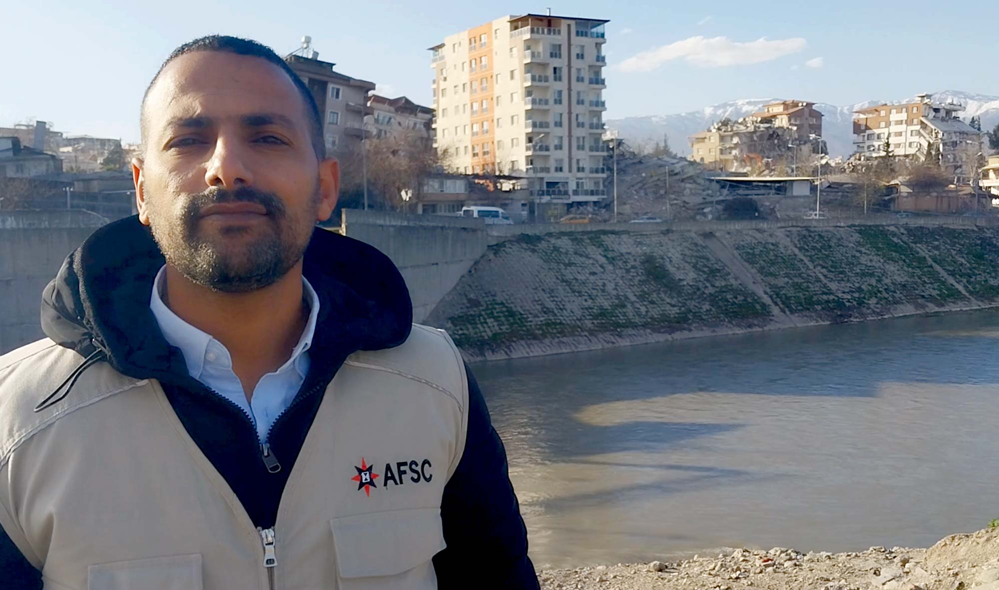 A man wearing an AFSC vest looks at the camera across a river from buildings damaged and destroyed by the earthquake