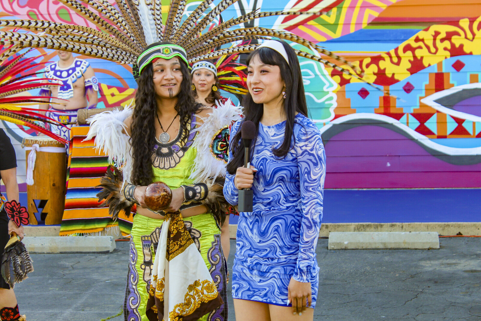 An artist speaking at a mural unveiling with dancers in indigenous costumes
