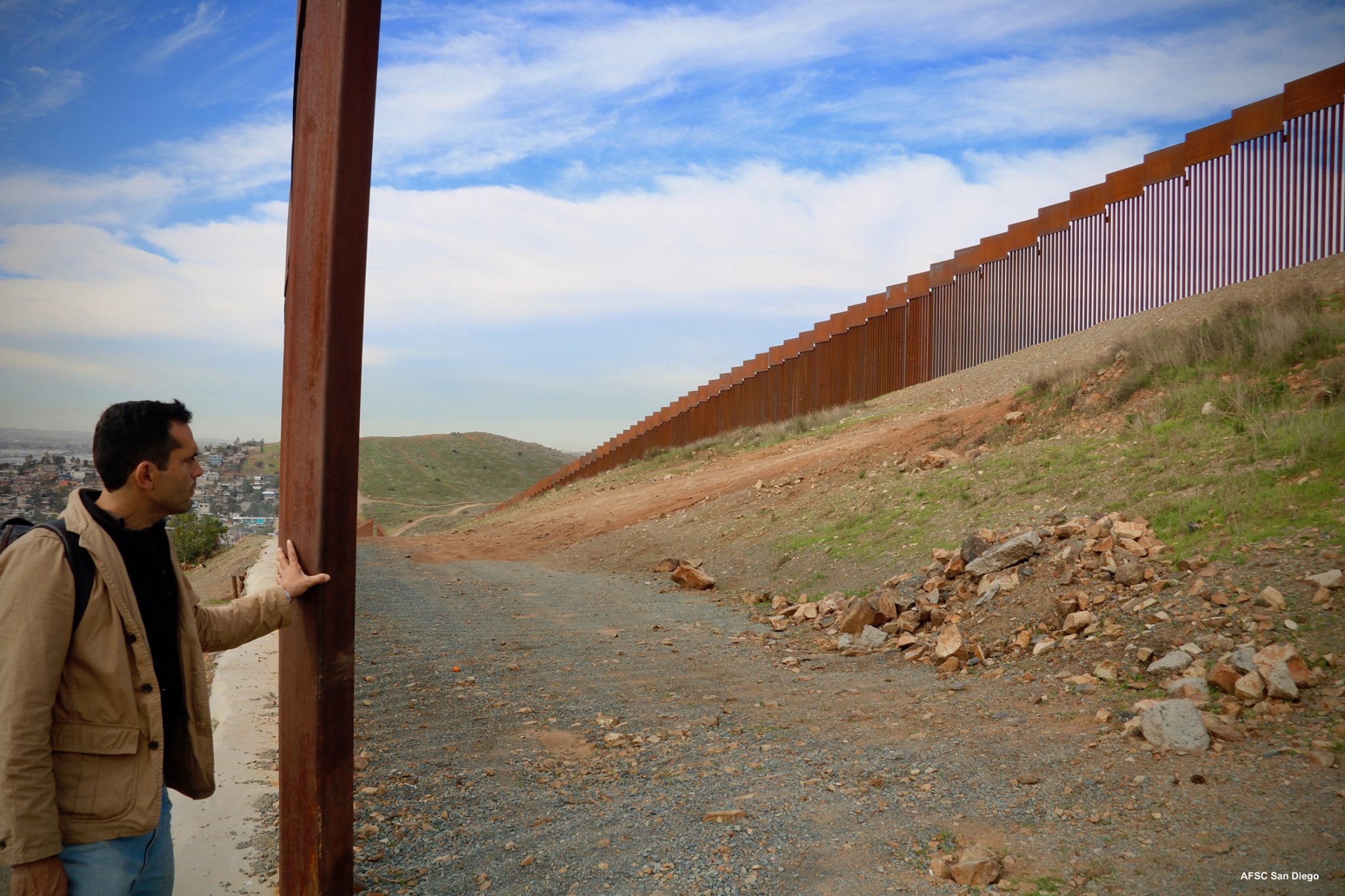 A man stands looking up at a tall fence along the border
