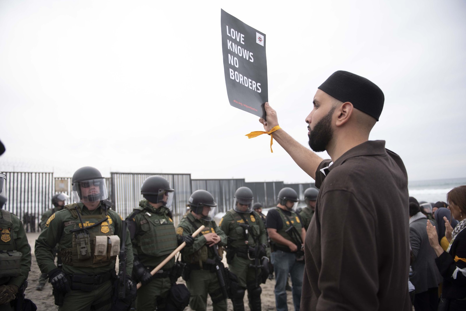Man holding a sign in front of armed guards at the U.S. Mexico border