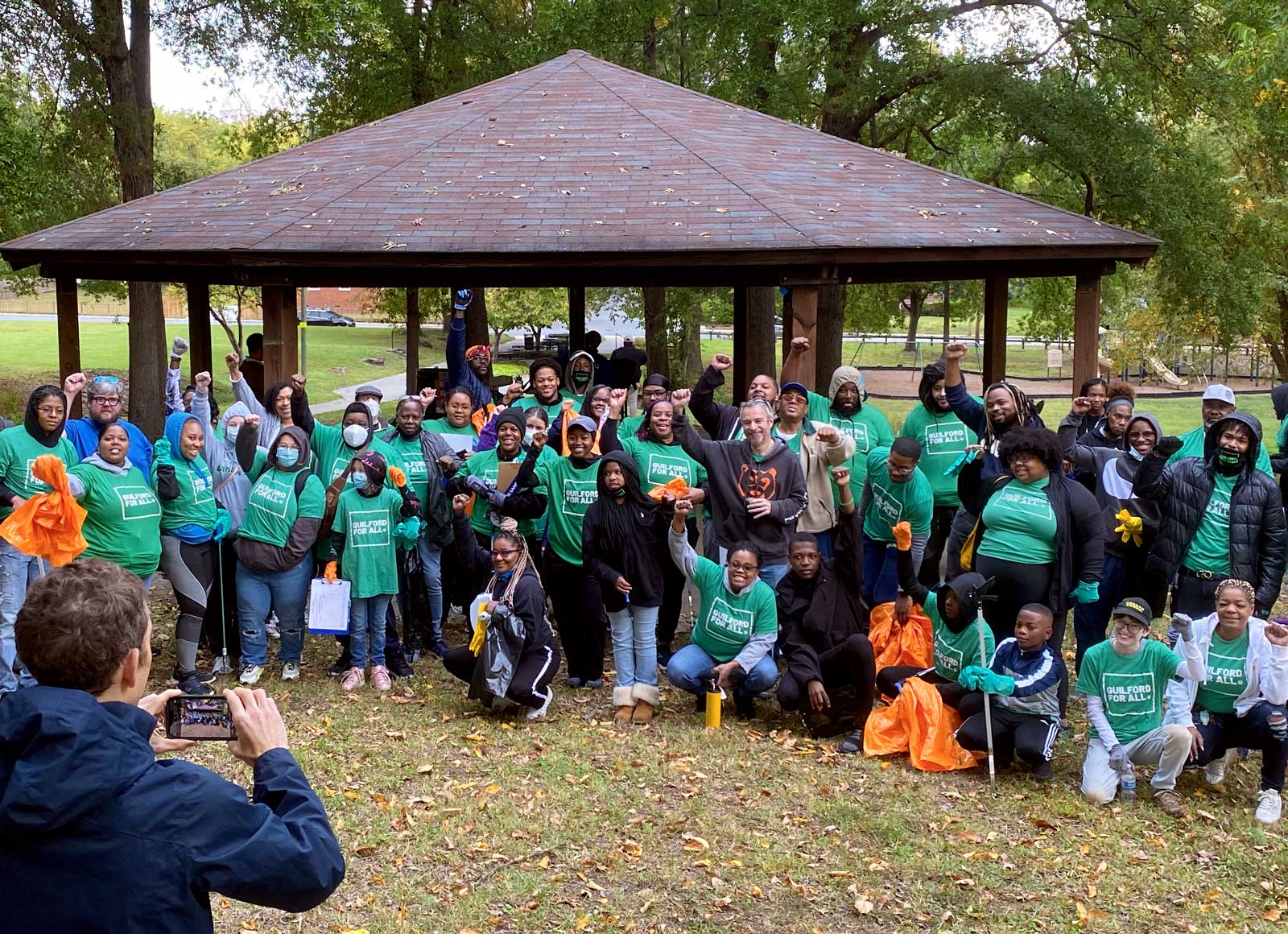 A group of community members with matching t-shirts smiles and raises fists in a park while a photographer takes their photo