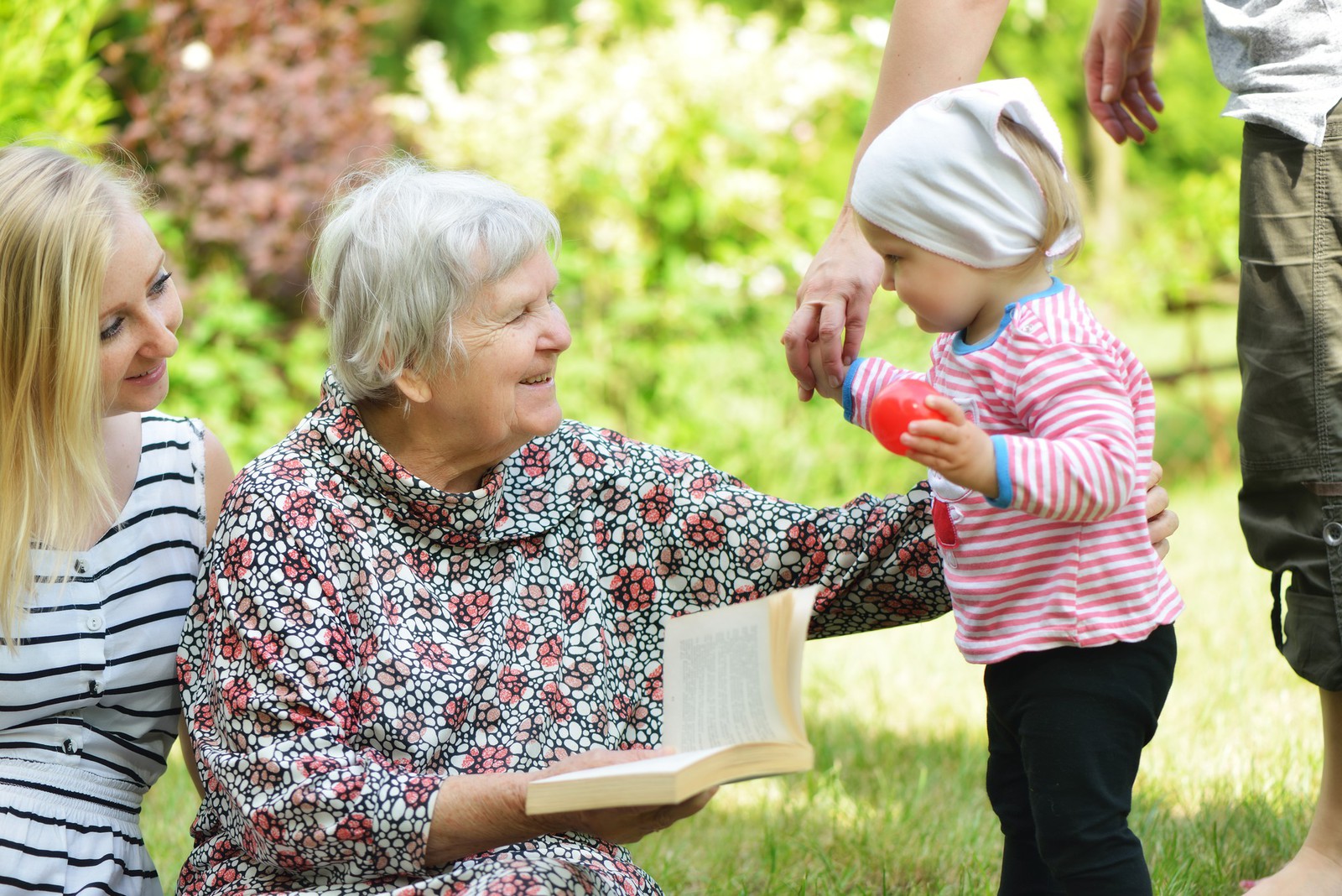 older woman holding book and smiling at toddler playing outside