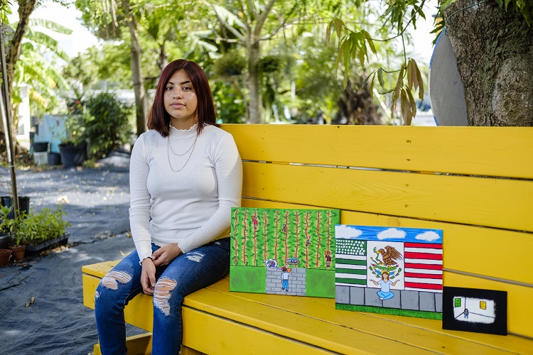 Photo of Alicia Villasenor sitting on a bench with three of their art pieces