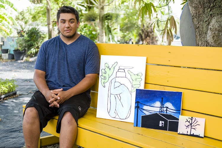 Photo of Bernard Villasenor sitting on a bench with three of their art pieces