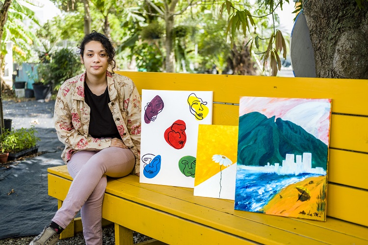 Photo of Ilse Moreno sitting on a bench with three of their art pieces