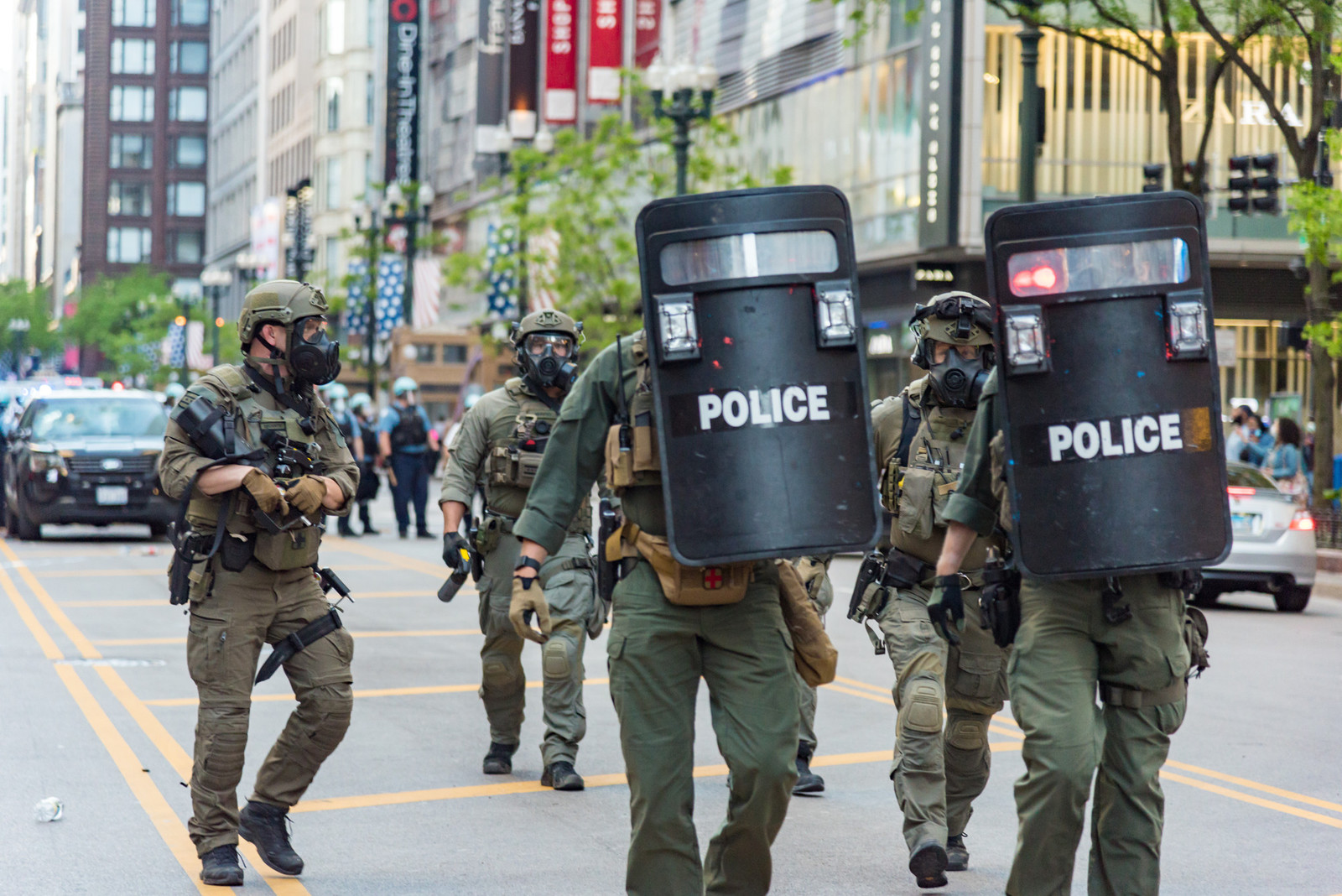 A group of militarized police walking down a city street, holding shields and riot gear.