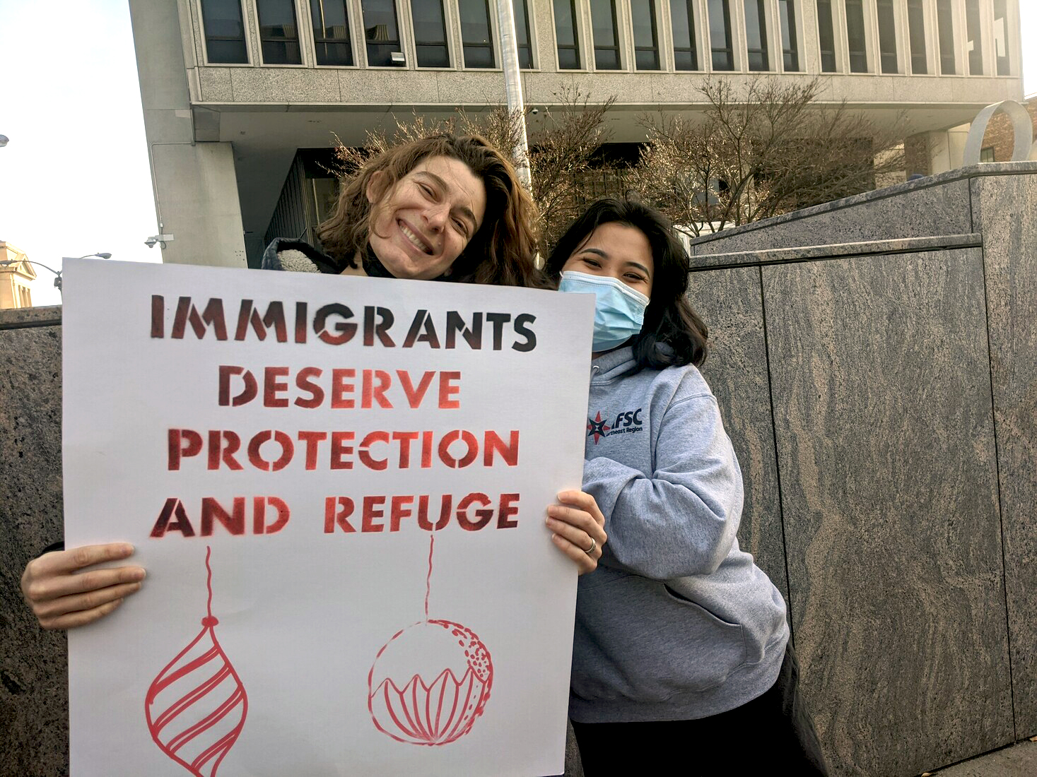 two people huddling outside and smiling holding sign that says immigrants deserve protection and refuge
