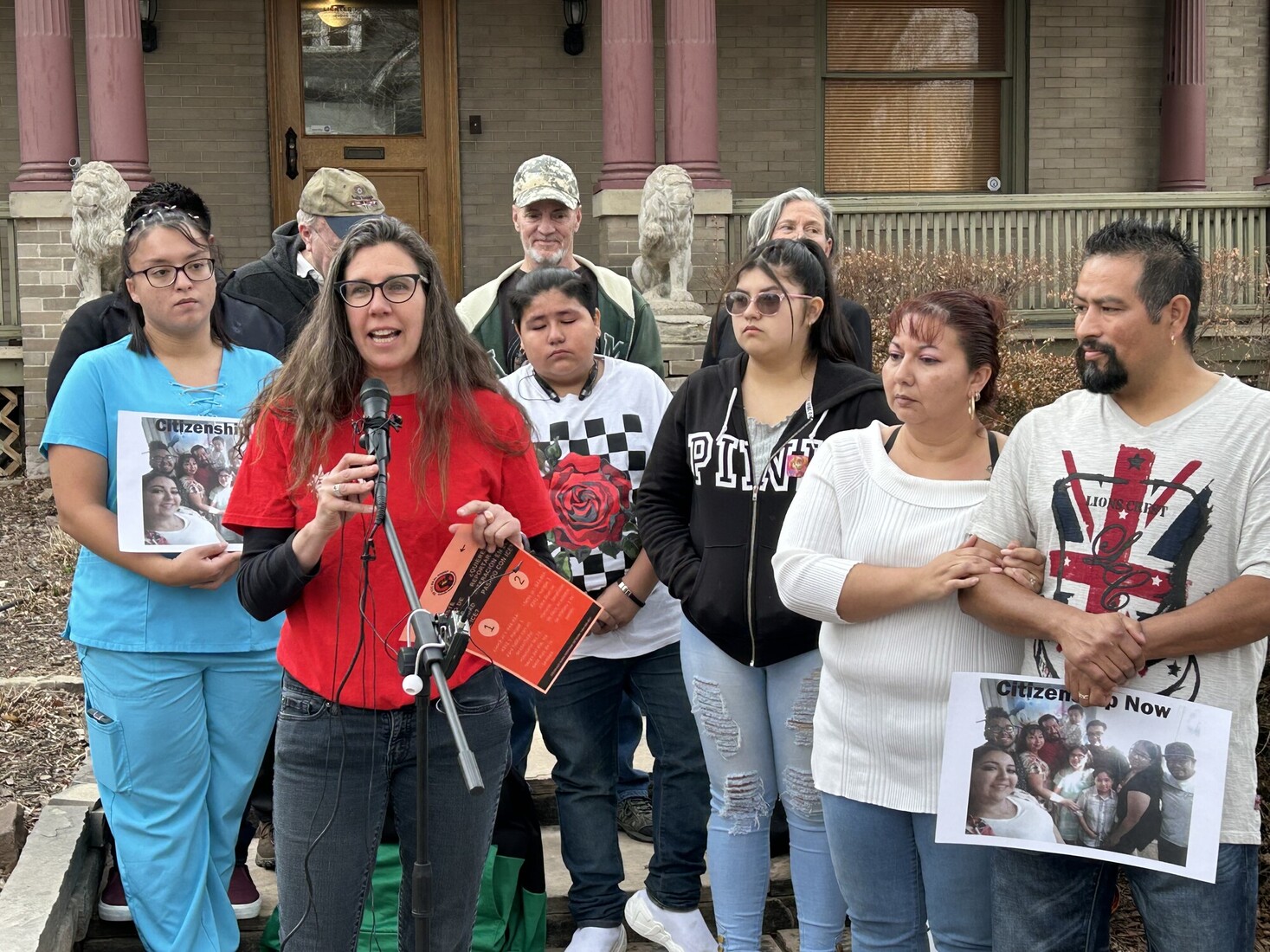 Woman in a red shirt speaking at a microphone outside a building, surrounded by a crowd