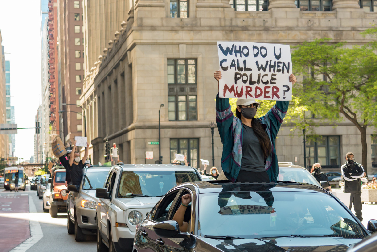 protester standing out the top of a car roof window, in a traffic protest. They are holding a sign that reads: who do you call when the police murder?