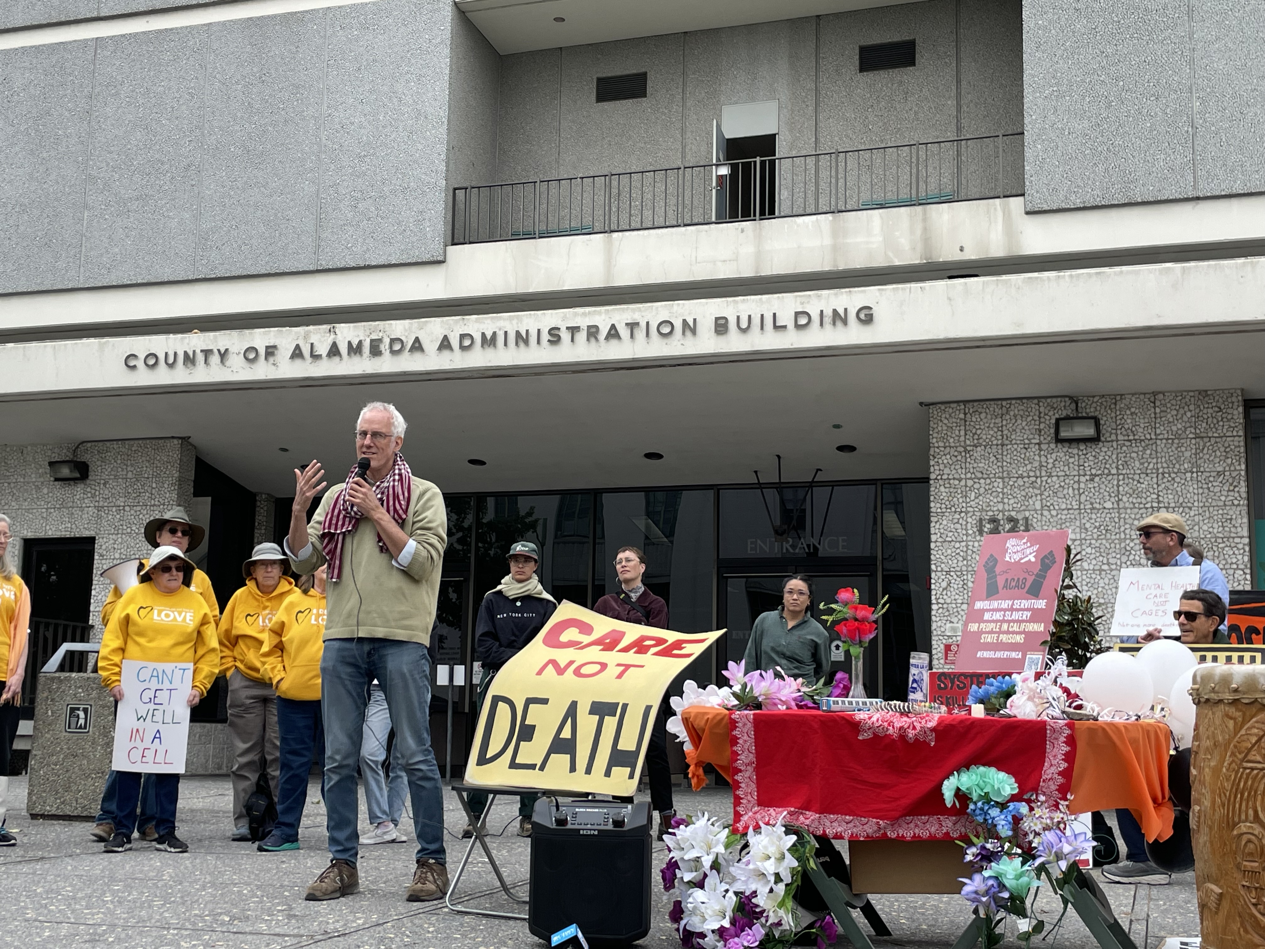 Photo of John Lindsay-Poland, California Healing Justice Program's Co-Director, addressing the crowd at the rally to call for halting Alameda County jail expansion, held in front of county building. He stands besides signs and an alter to those who've lost their lives at the jail.