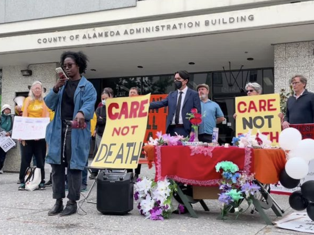 Photo at a rally. A woman reads names and pours libations from her water bottle. She stands in front of an alter and signs calling for CARE NOT DEATH.