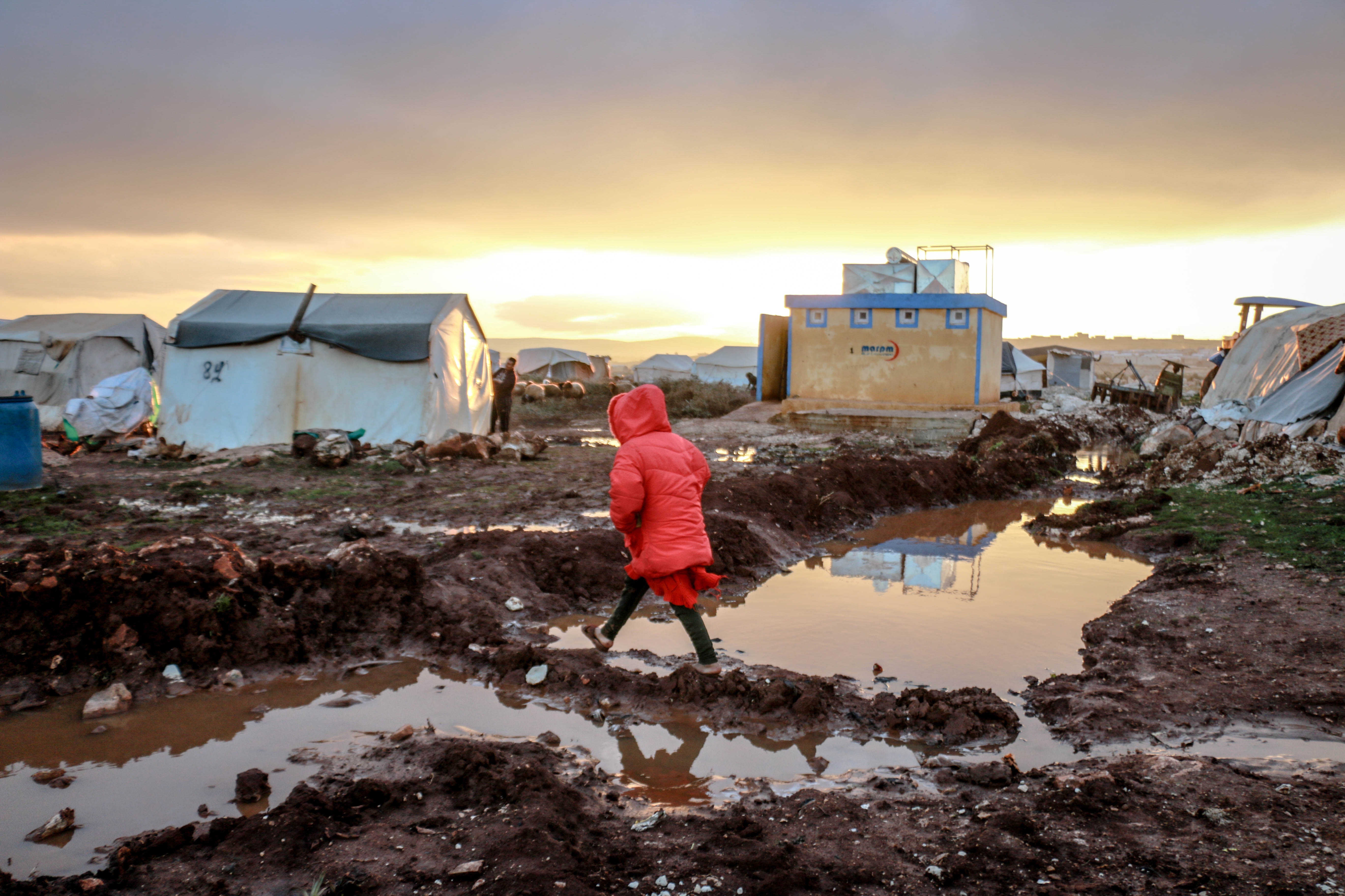 child in coat walking across muddy field with tents in the background