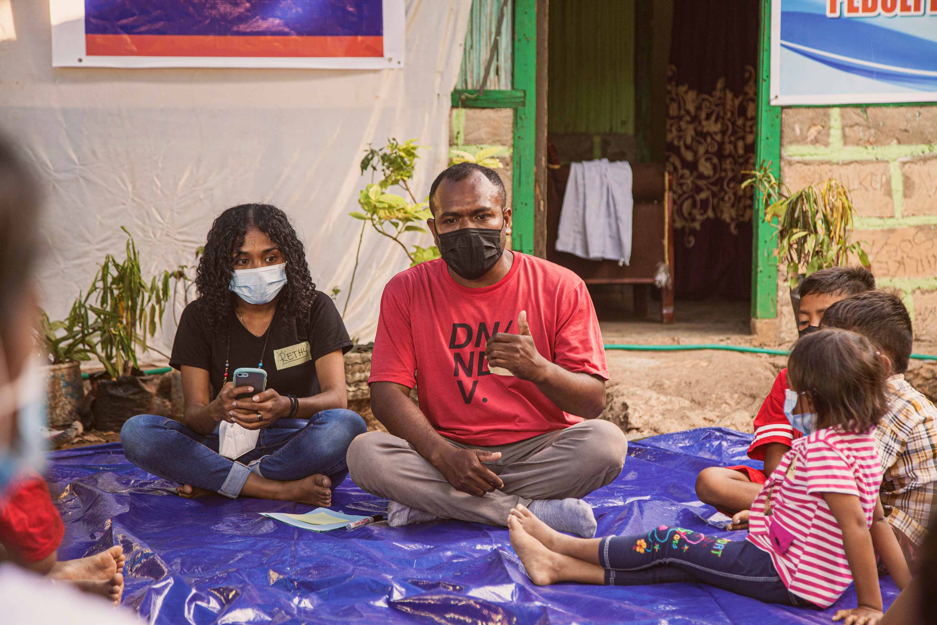 Two adults sitting outoors on the ground with several small children in Indonesia