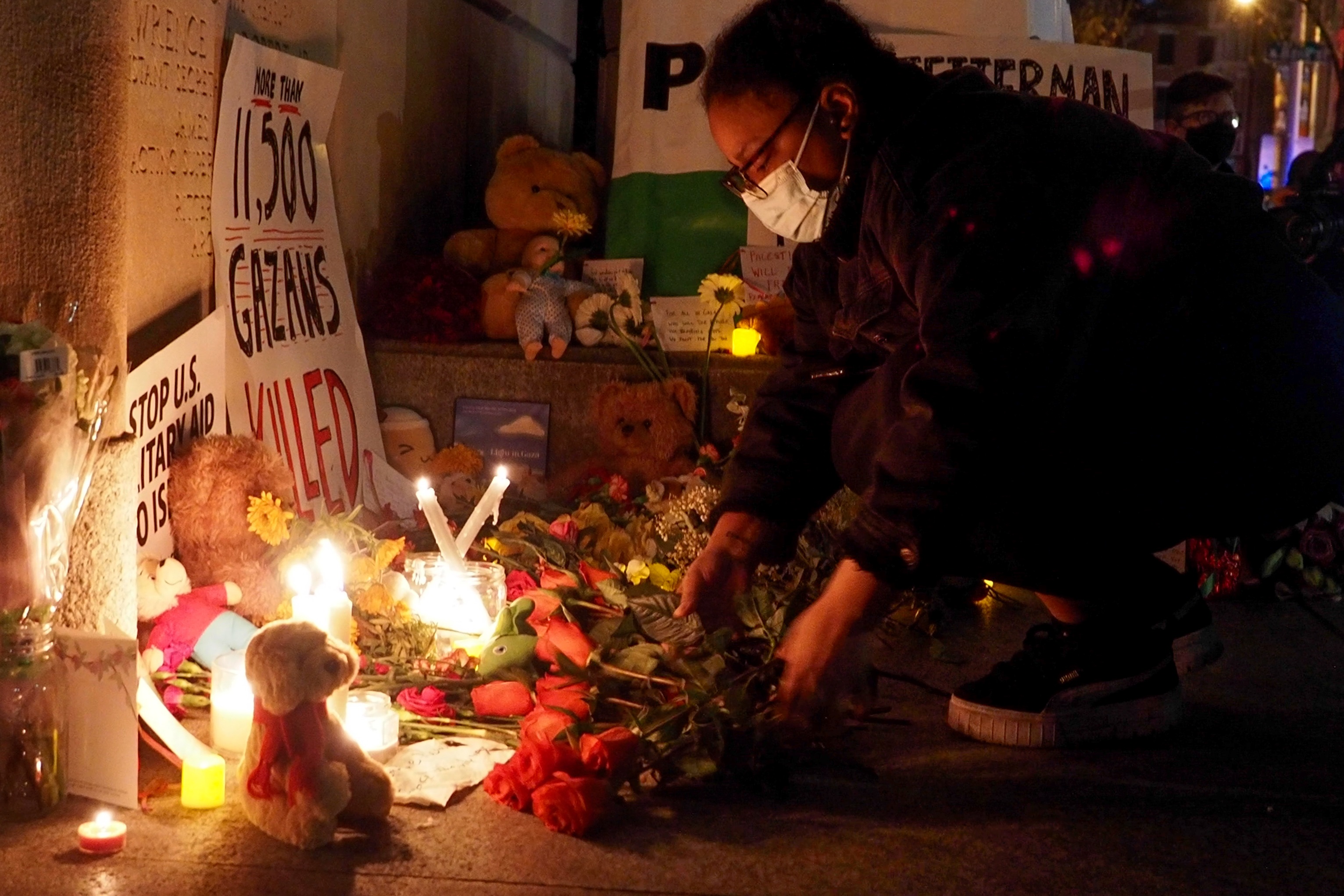 Person with mask kneels on ground in front candles, flowers, and signs