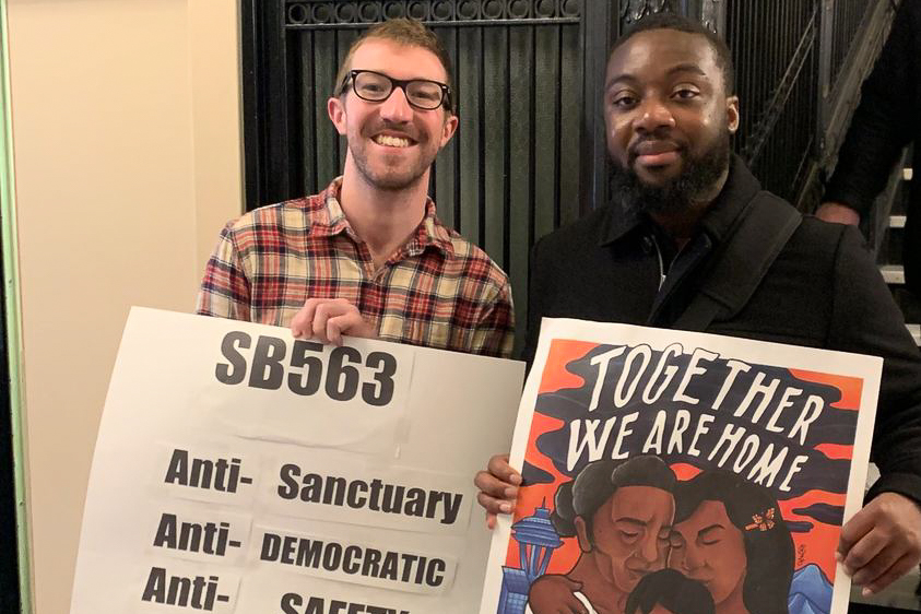 Image of two people holding signs that say 'SB563' and 'Together we are home'