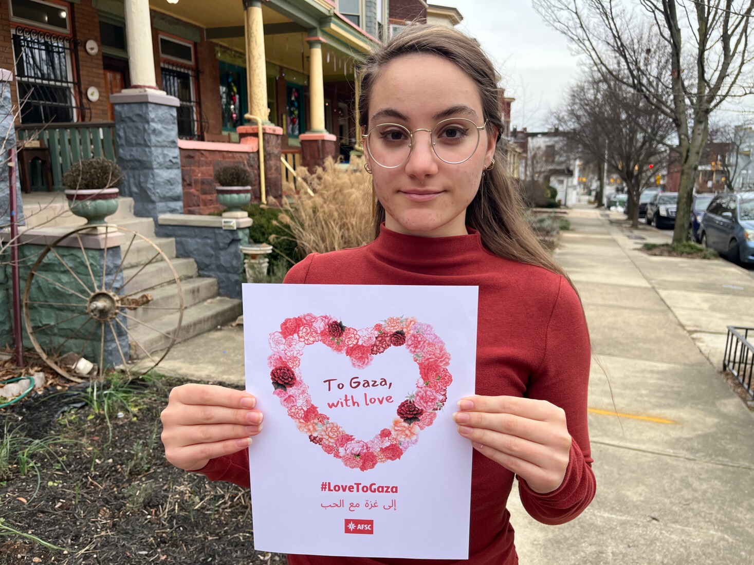 Person stands on sidewalk holding sign with a heart that reads 