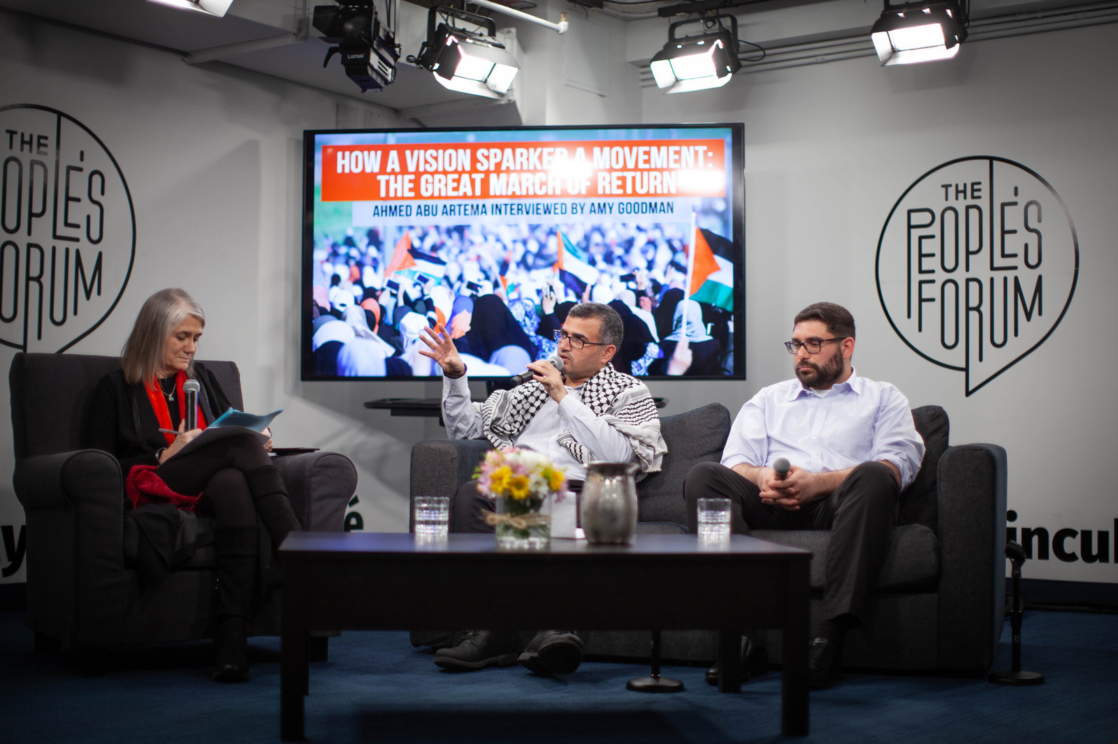 Three people speaking on a stage in front of a large screen.