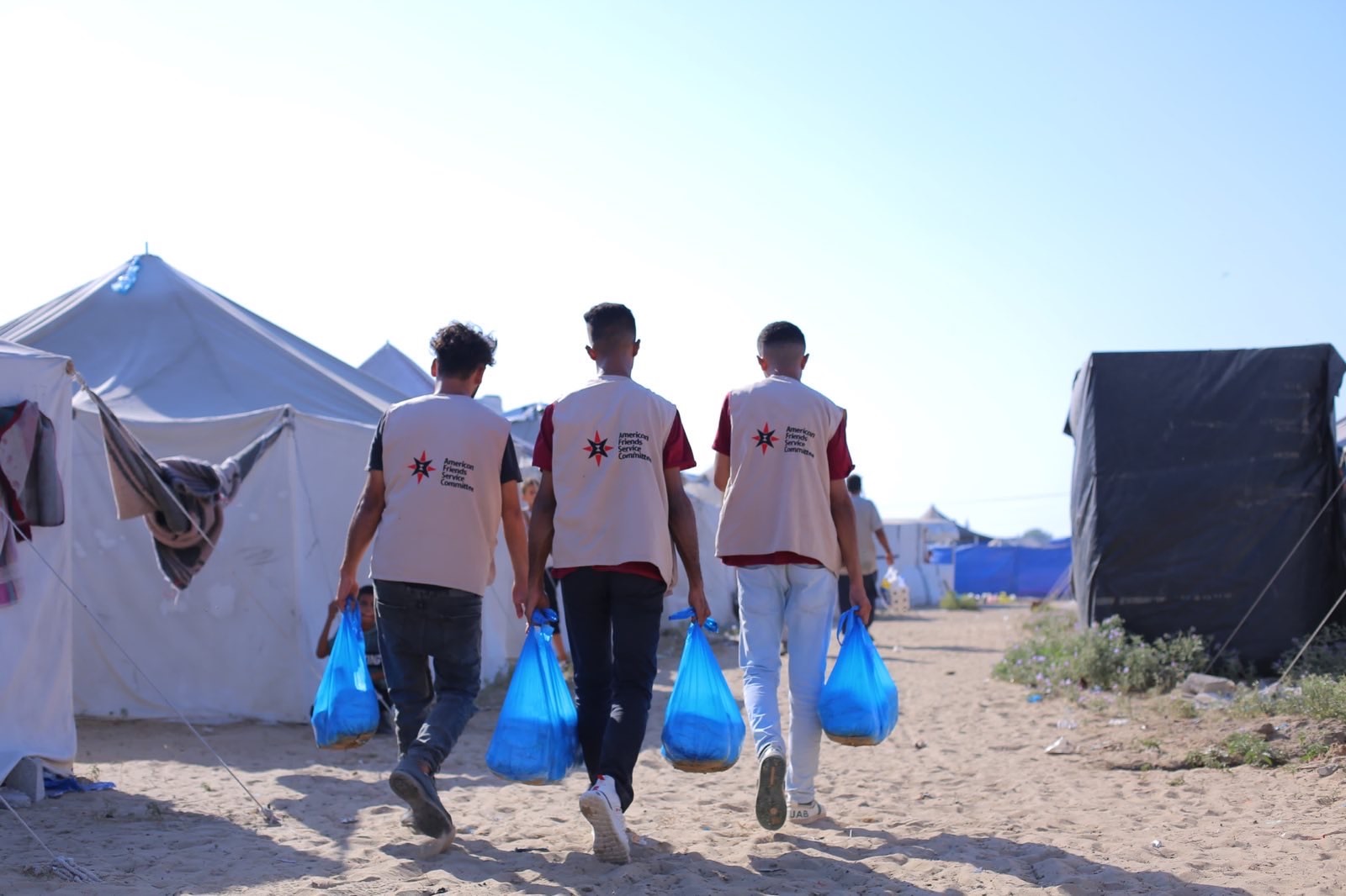 3 men with AFSC vests walk among tents holding blue plastic bags