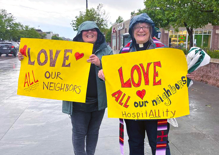 Two people holding protest signs at a rally: Love all our neighbors