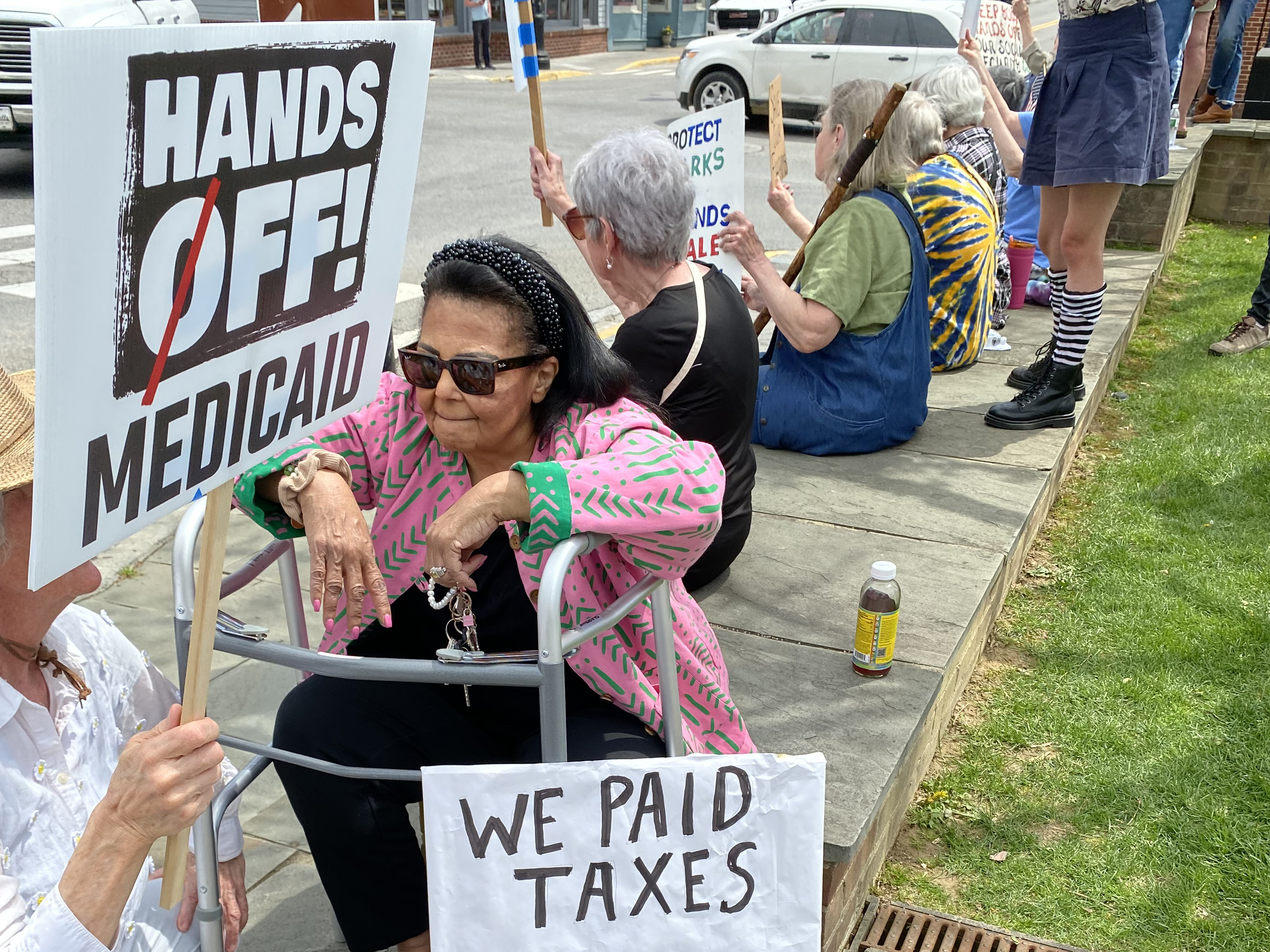 Image of a woman with a walker behind a sign that says 'Hands off! Medicaid'