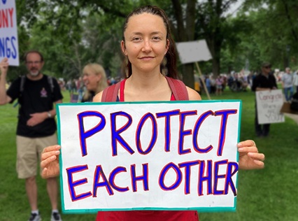 Young person holding a handwritten sign in bold colors: Protect each other