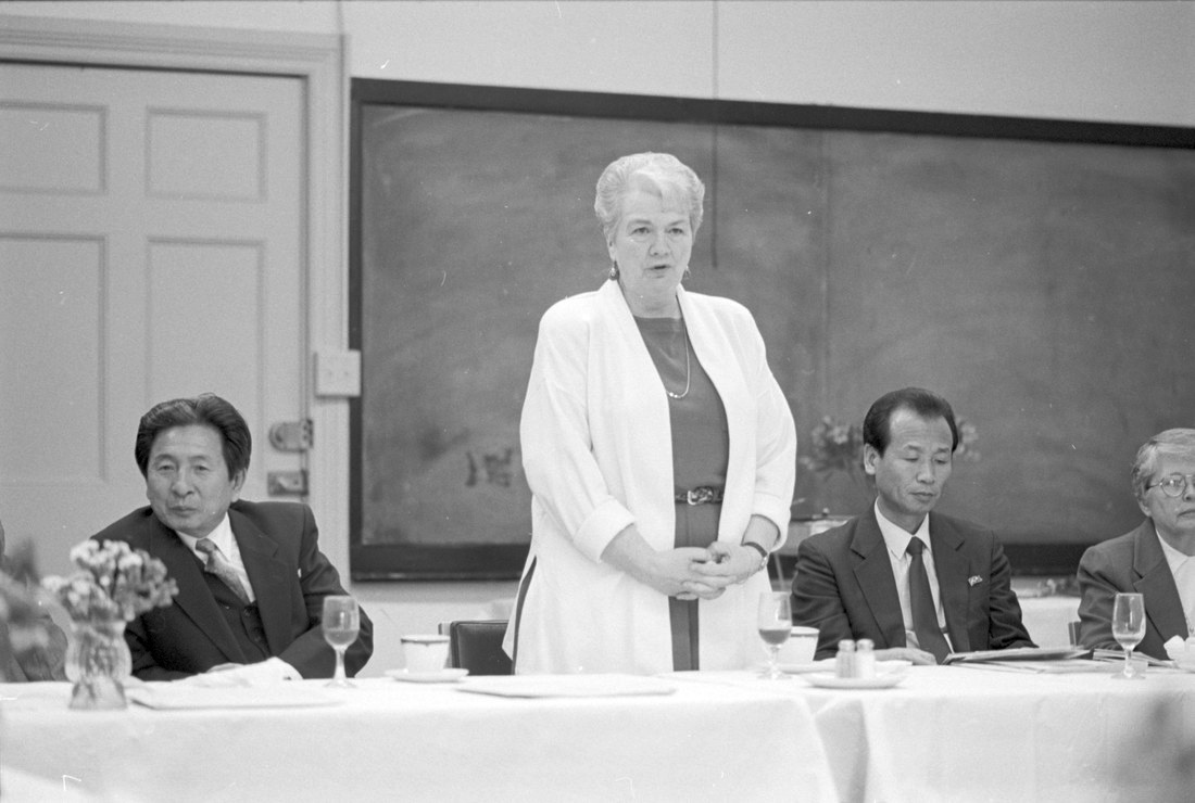 four people at a long table, the woman in the middle is standing up to speak