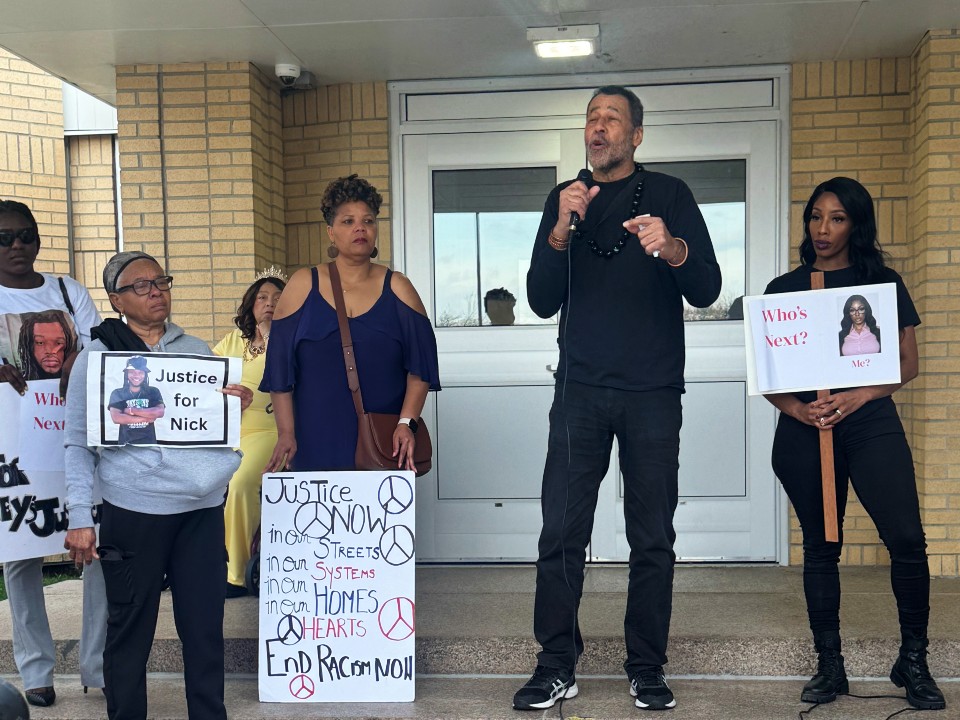 A man speaks on a mic in front of a house with 5 others around him holding signs