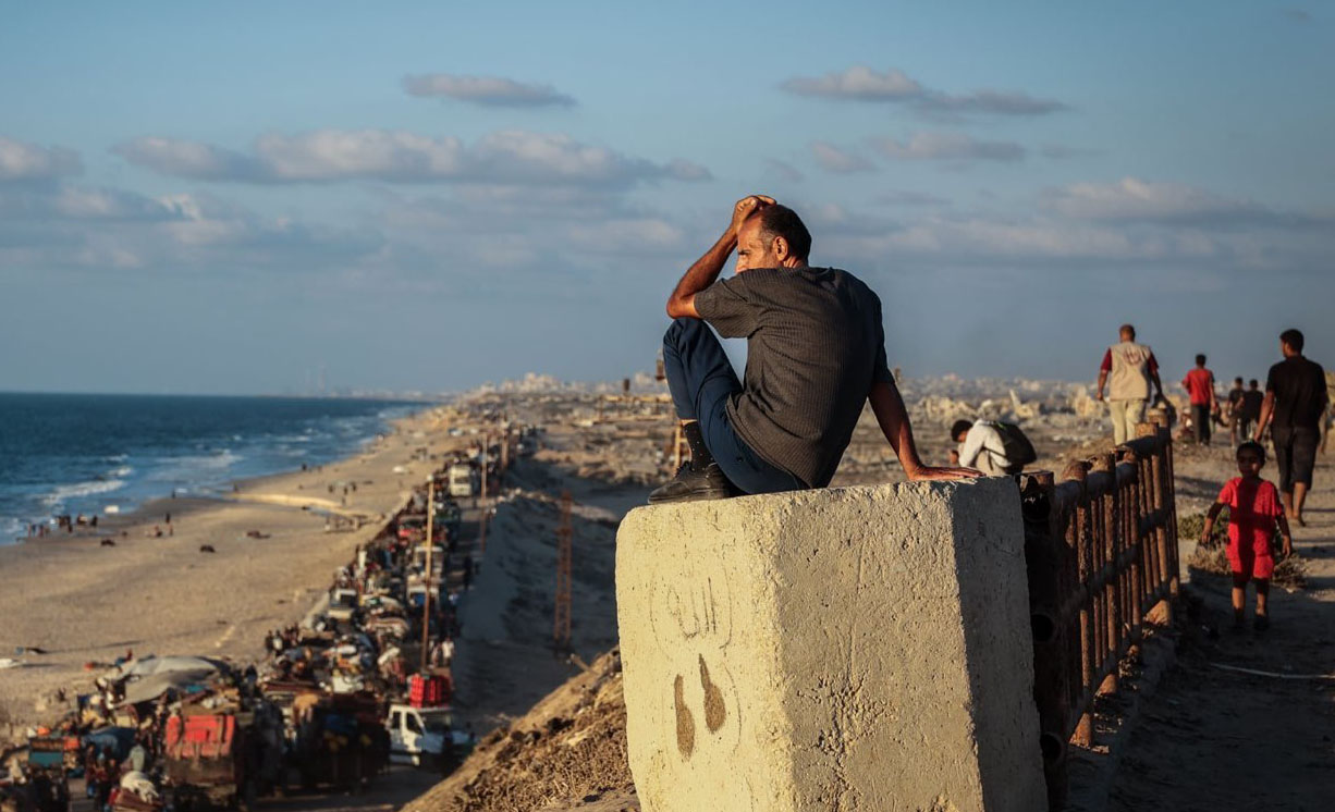 man looking saddened sitting on top of concrete block. He is looking out into the sea, other displaced people are taking refuge on the beach. 