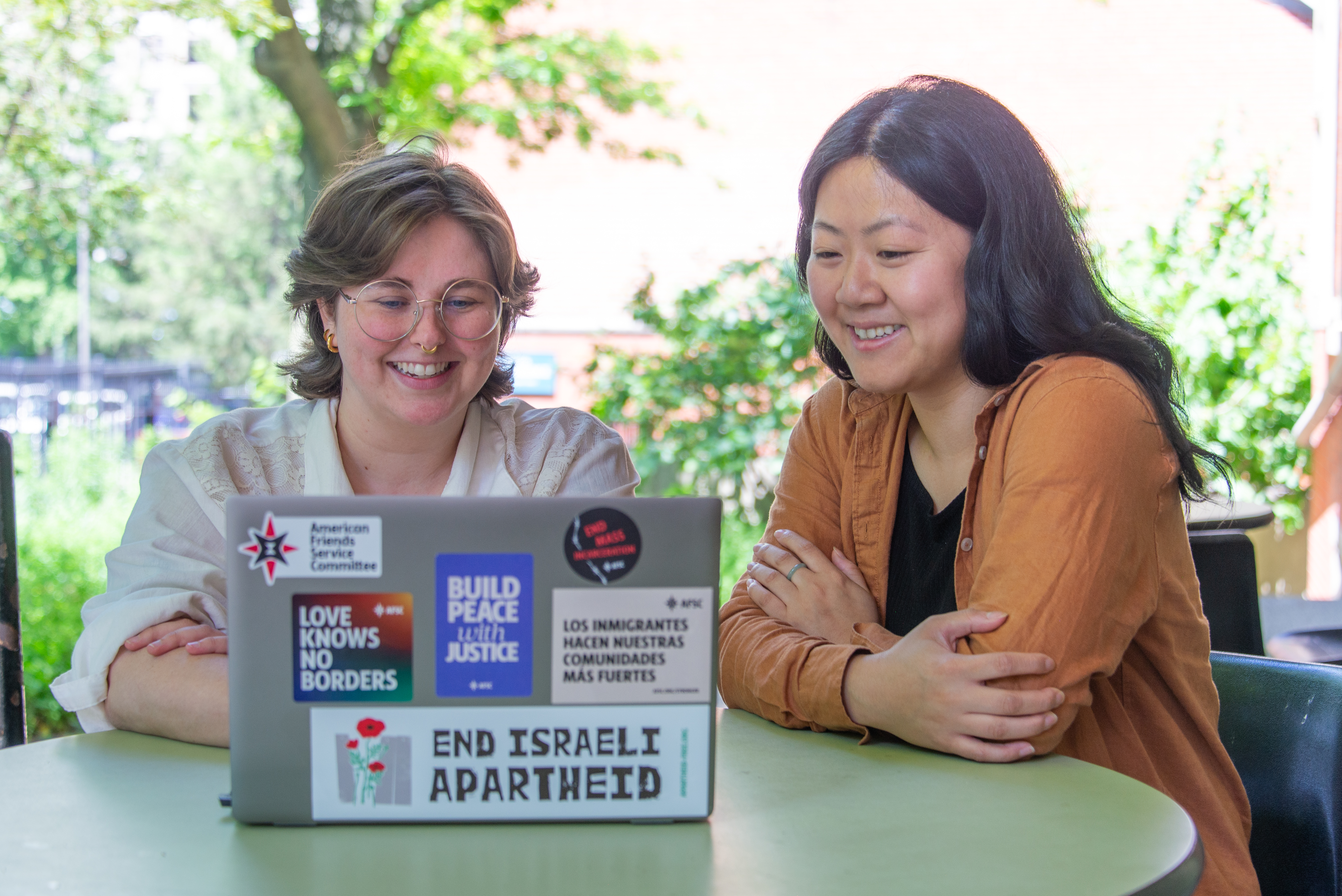 two people sitting at a table outside smiling and looking at a laptop. the laptop has many protest stickers on it, one reads 