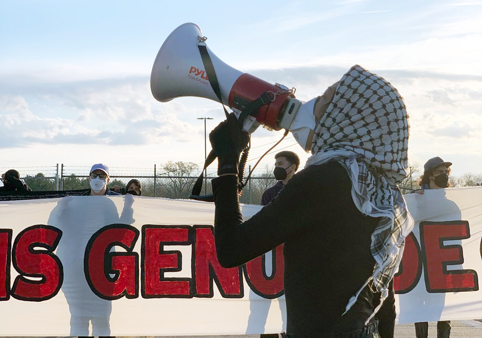 person in kaffiyeh with a megaphone. behind is a banner that reads 